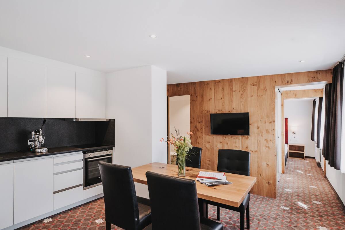 Une cuisine moderne avec des armoires blanches, un comptoir noir et un four encastré. On y trouve un coin repas avec une table en bois et des chaises noires, ainsi qu’un téléviseur fixé au mur.
