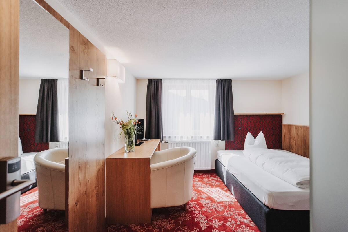 Une chambre d’hôtel comprenant un lit, un bureau avec une chaise, une petite salle de bain et une fenêtre avec des rideaux.