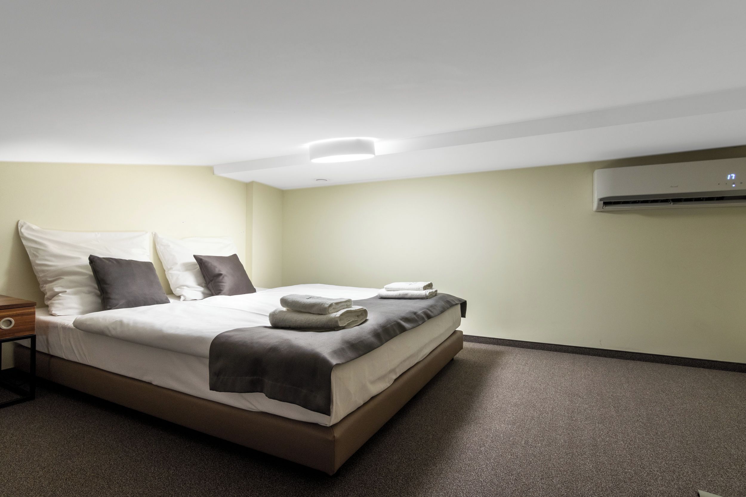 A neatly made bed with pillows and a folded towel, a bedside table, and an air conditioning unit on the wall.