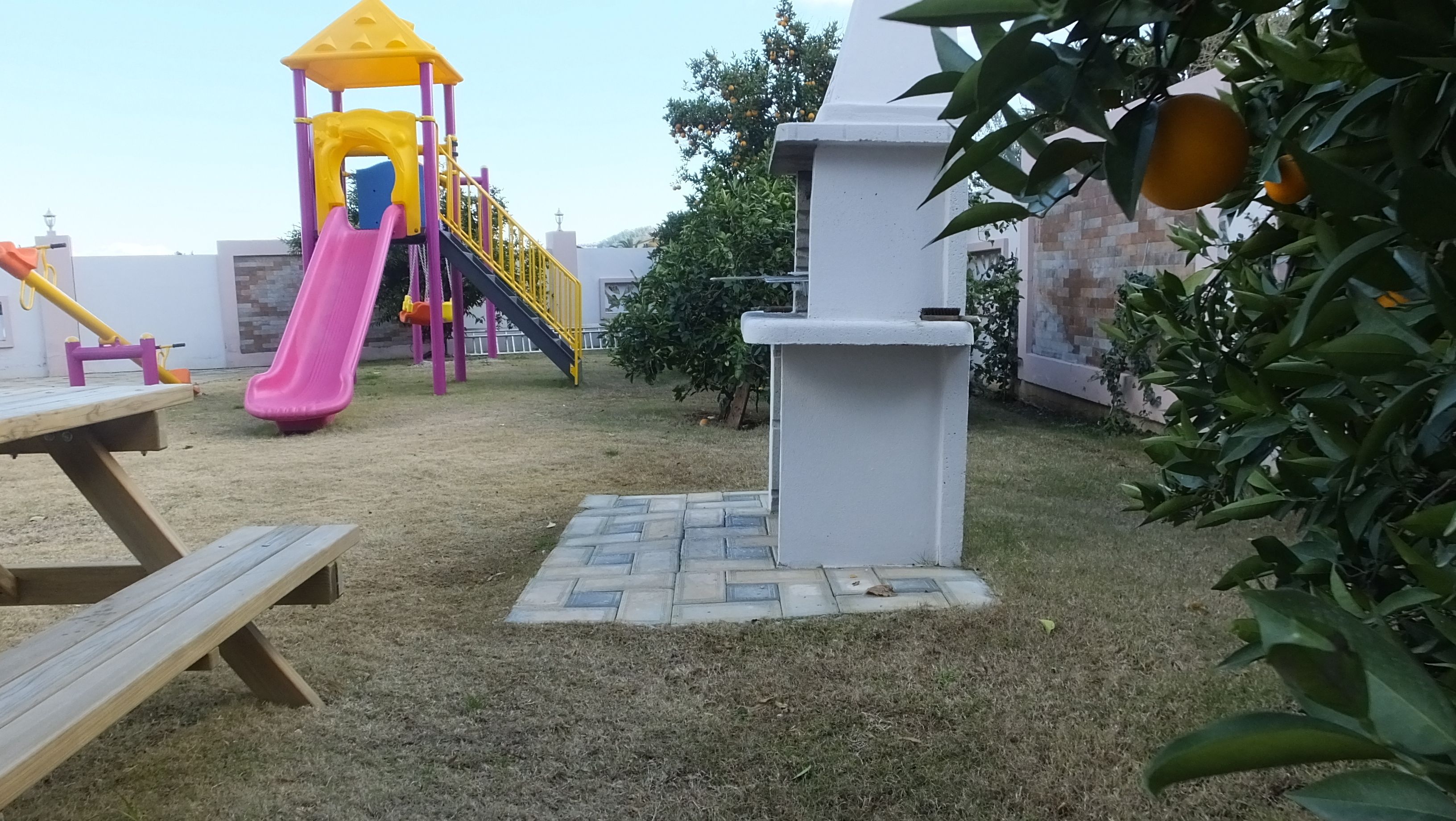 A playground with a slide and climbing structures, surrounded by a grassy area and a white wall.