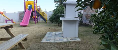 A playground with a slide and climbing structures, surrounded by a grassy area and a white wall.