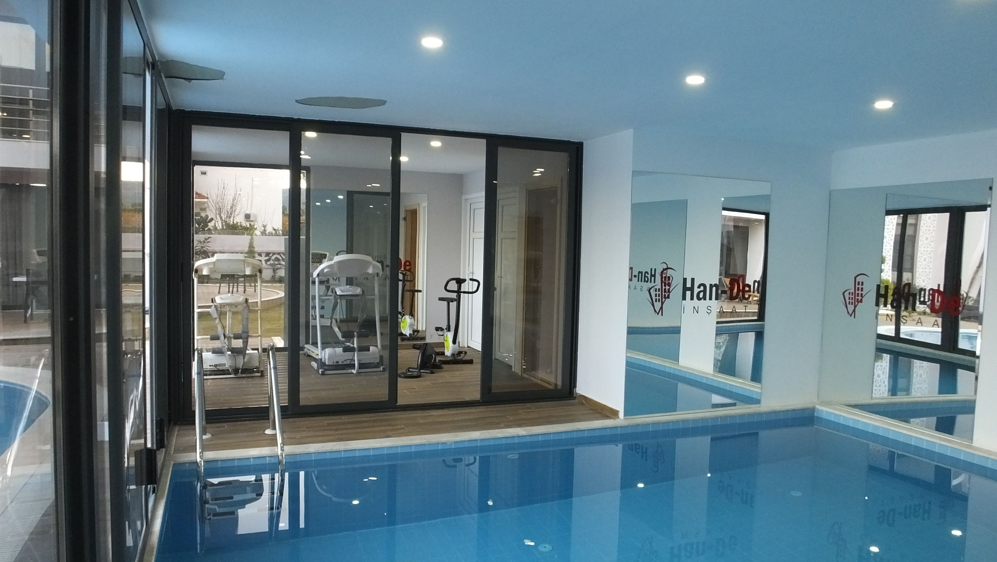 A modern indoor swimming pool with glass walls, exercise equipment, and a visible logo for 'Han-Den'.