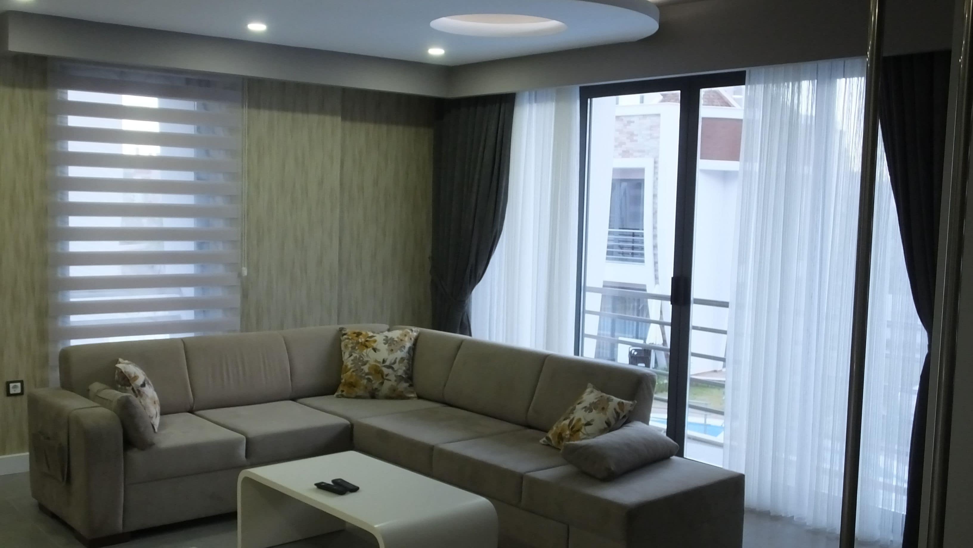 Una sala de estar moderna con un sofá modular, una mesa de centro blanca y ventanas grandes con persianas y cortinas.
