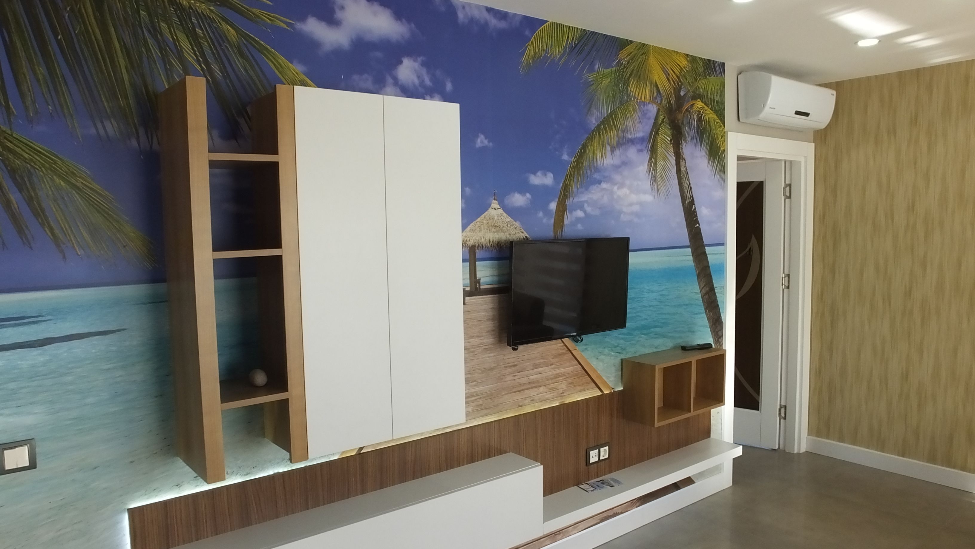 A room with a TV mounted on a wall with a tropical beach mural, wooden furniture, and a built-in air conditioning unit.