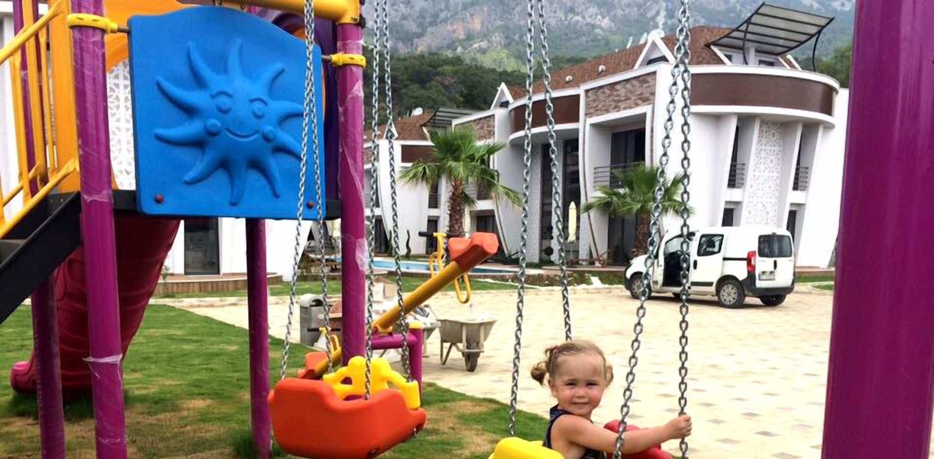 A playground with swings and a slide, set against a backdrop of a building with a white facade and a vehicle parked nearby.