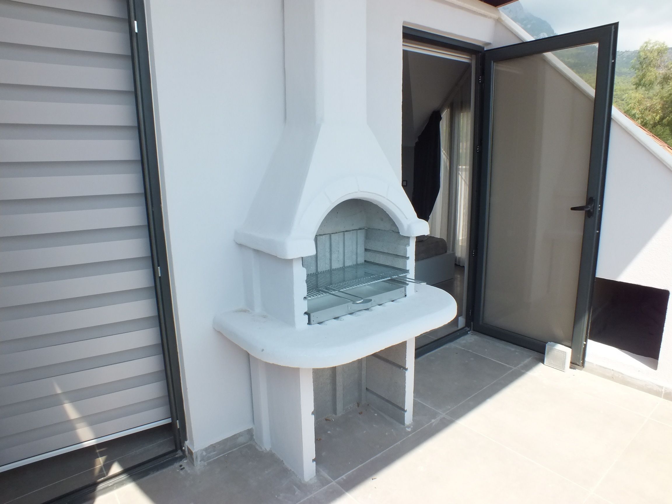 A white outdoor pizza oven with a metal shelf, located on a balcony with a sliding glass door and a view of greenery.
