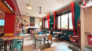 Interior de restaurante con varias mesas y sillas, una barra donde una persona atiende, y un amplio ventanal con cortinas.
