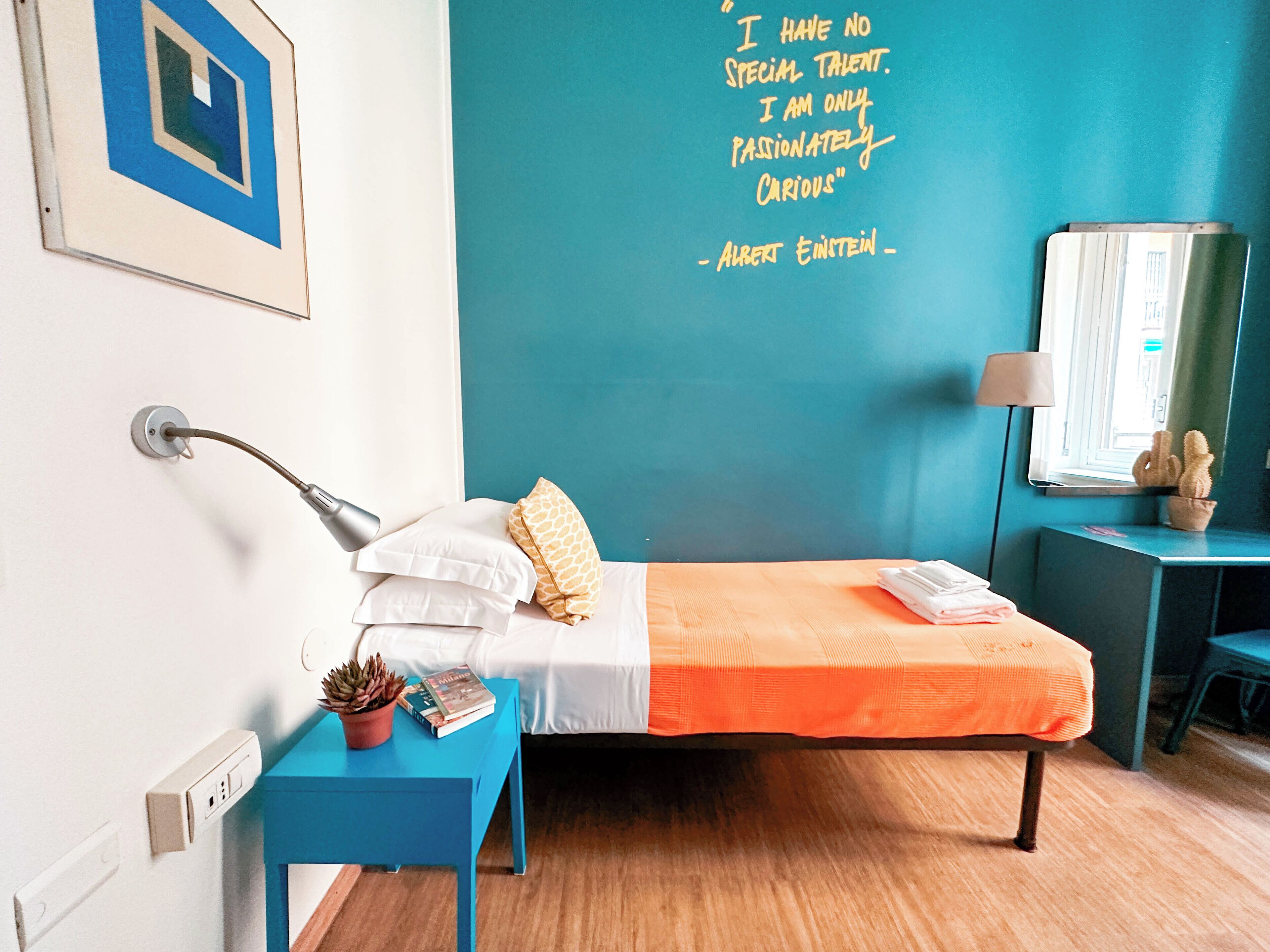 A neatly arranged hotel room with a bed, a bedside table with a lamp, a small potted plant, and a wall with a motivational quote.