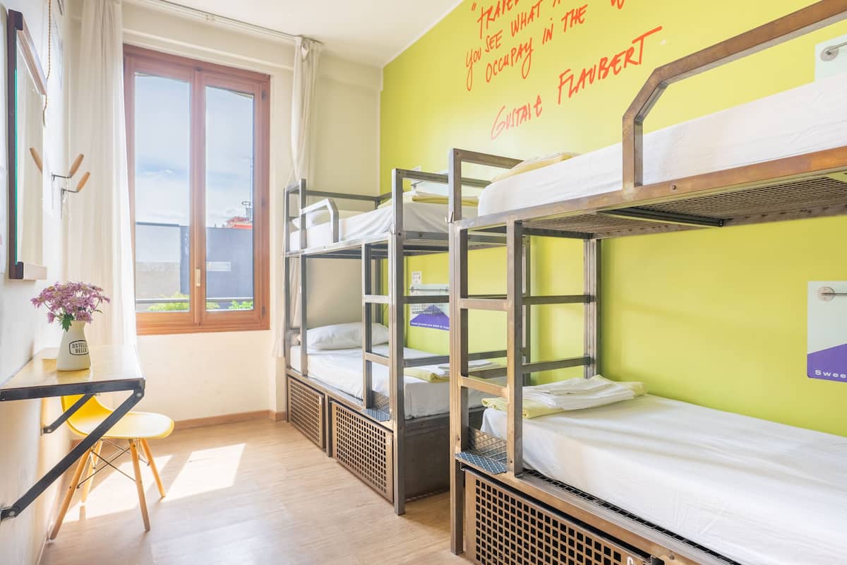 A dormitory room with bunk beds, a desk with a chair, a window with curtains, and a wall with a motivational quote.