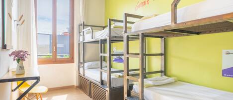 A dormitory room with bunk beds, a desk with a chair, a window with curtains, and a wall with a motivational quote.