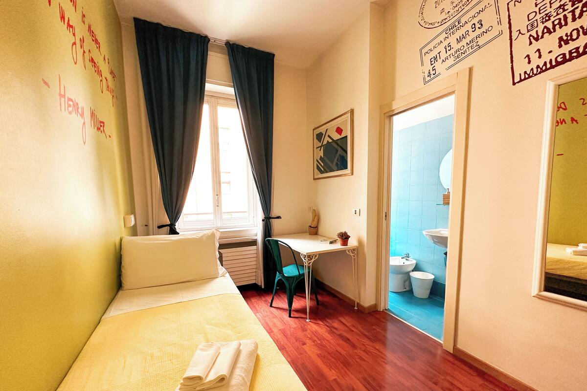 Habitación de hotel con cama, escritorio, silla, ventana con cortinas, lavamanos y un inodoro.