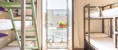 A dormitory room with bunk beds, a ladder, a small table with a plant, and a view through a glass door.