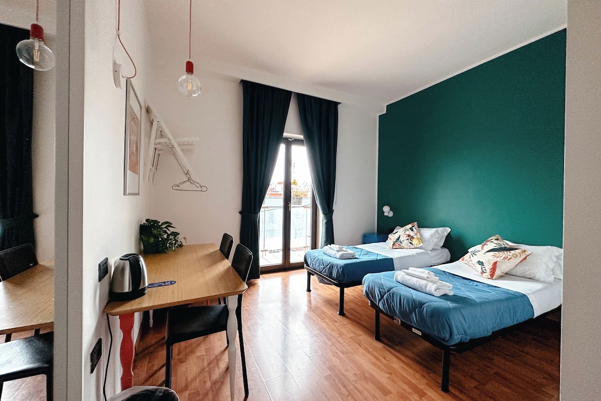 Habitación con una mesa de madera, dos camas con ropa de cama azul, una lámpara colgante y vistas a través de una ventana con cortinas.