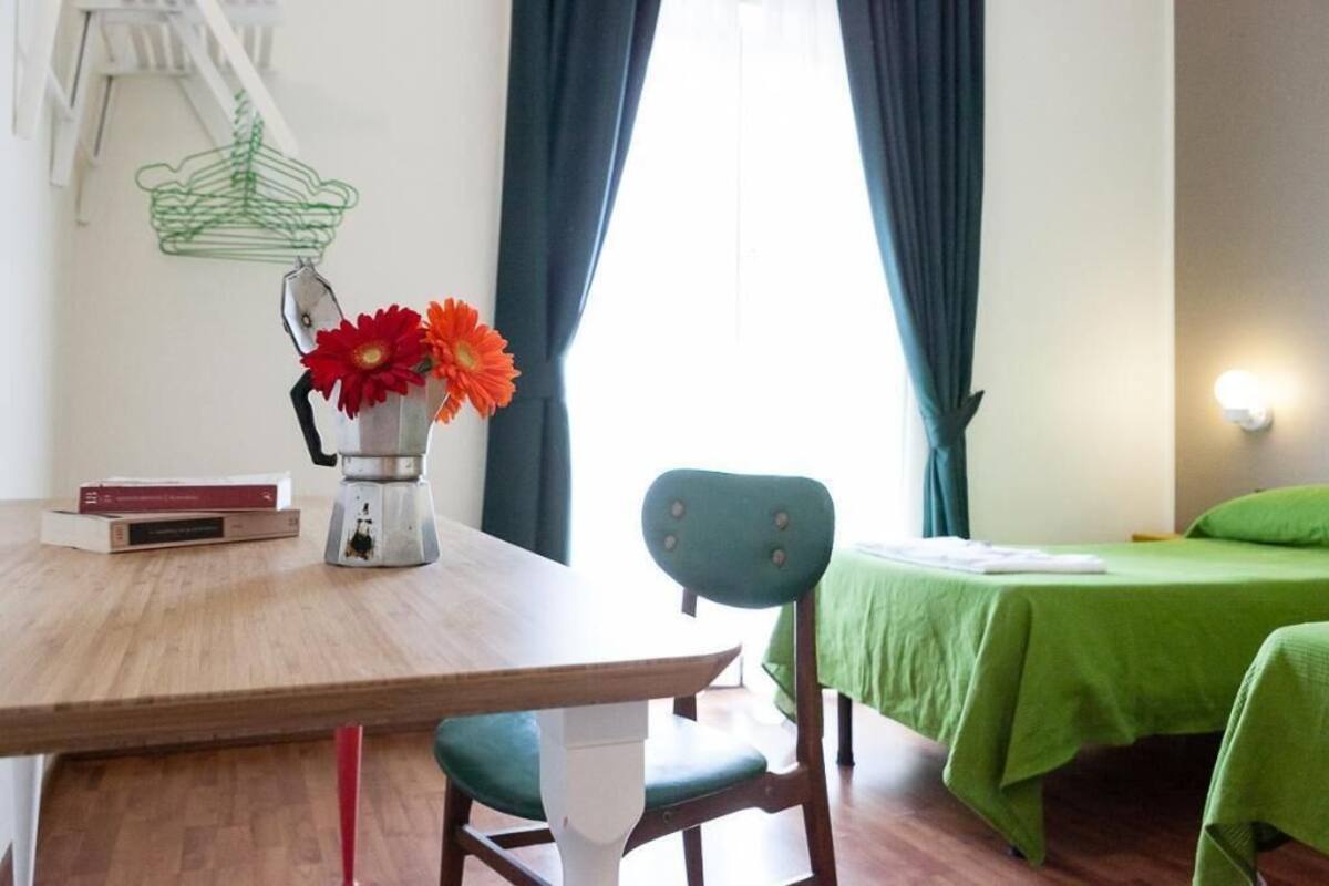 Una habitación con una mesa de madera, una silla, una cama con colcha verde, otra silla verde, una lámpara blanca y un adorno de perchero de metal verde.