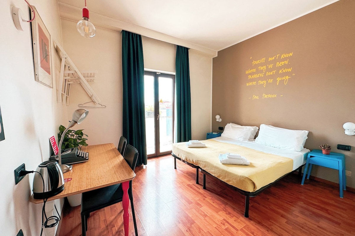 Una habitación de hotel con cama, mesitas de noche, escritorio con tetera, silla, planta en maceta y una pared con una cita.