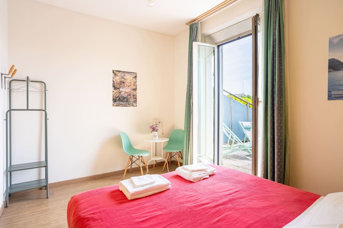 A hotel room with a bed, a small table, two chairs, a window with a view, and a metal shelf.