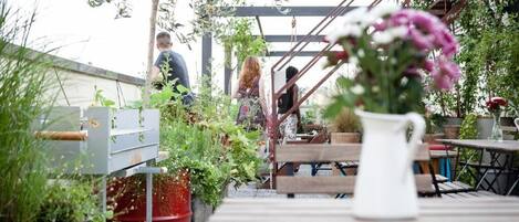 An outdoor patio area with wooden tables and benches, surrounded by potted plants and a metal staircase.