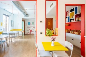 A brightly lit room with tables and chairs, a yellow table with a vase, and a red wall with shelves. - Ostello Bello Milano Centrale (Milan)