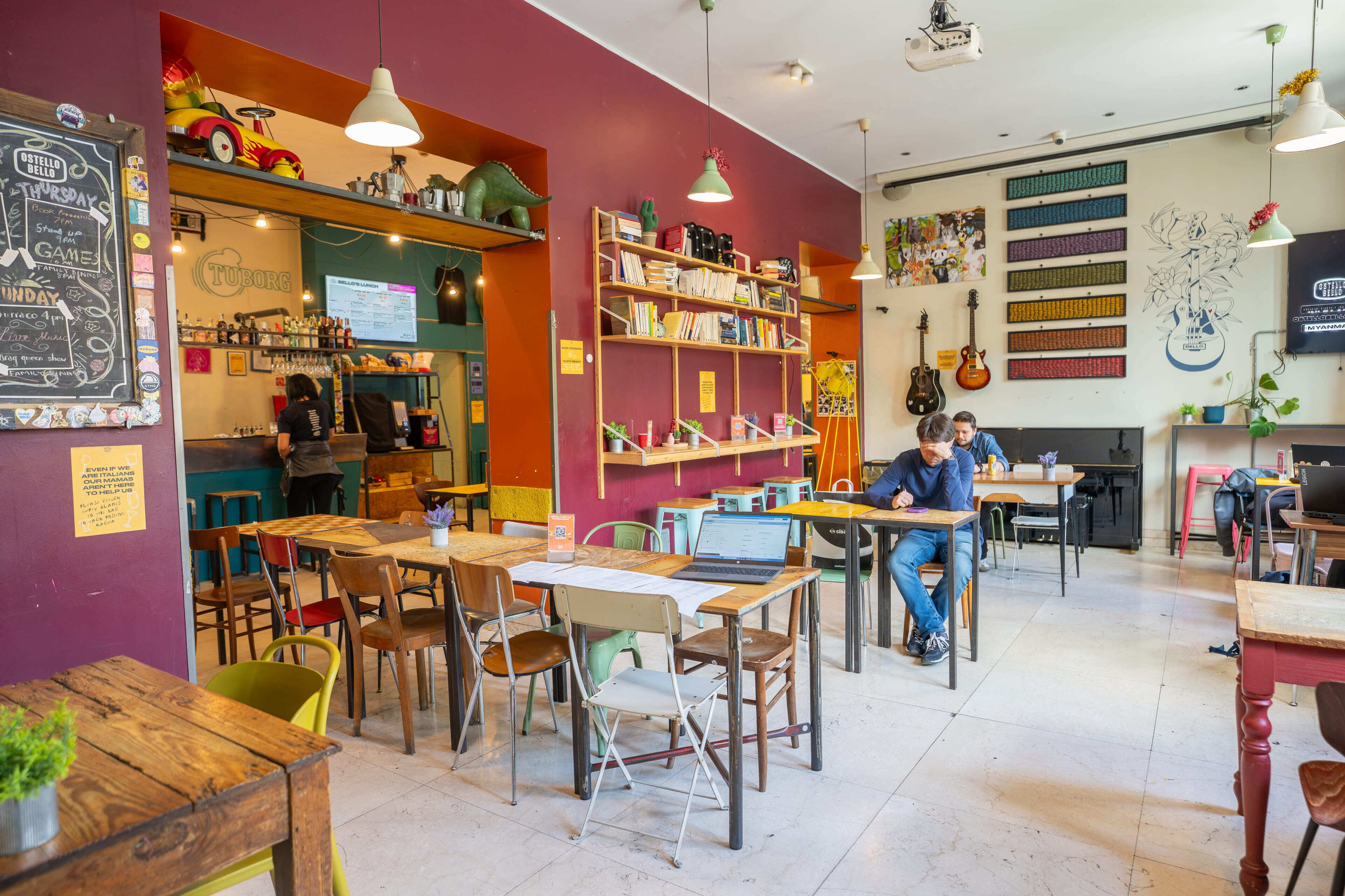 Un café con mesas y sillas, un menú en pizarra y estanterías con libros y plantas.