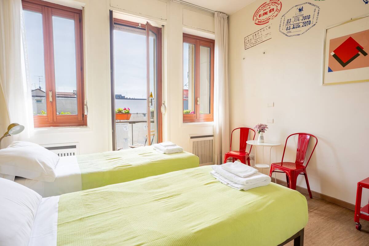 A hotel room with two beds, a small table with a chair, a radiator, and a window with a view.
