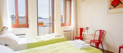 A hotel room with two beds, a small table with a chair, a radiator, and a window with a view.