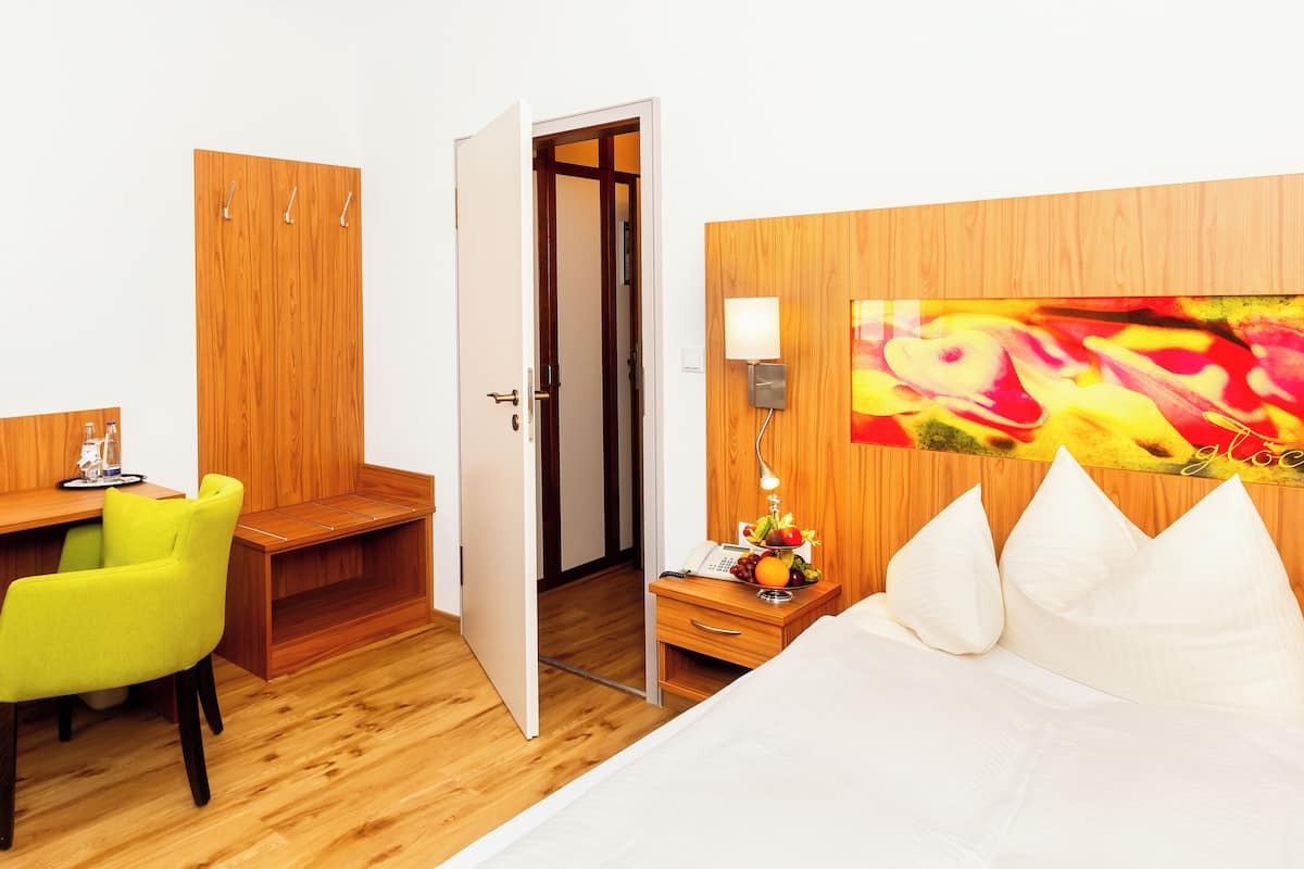 A hotel room with a wooden desk, a bright green chair, a bed with white pillows, a nightstand with a lamp, and a colorful abstract painting on the wall.