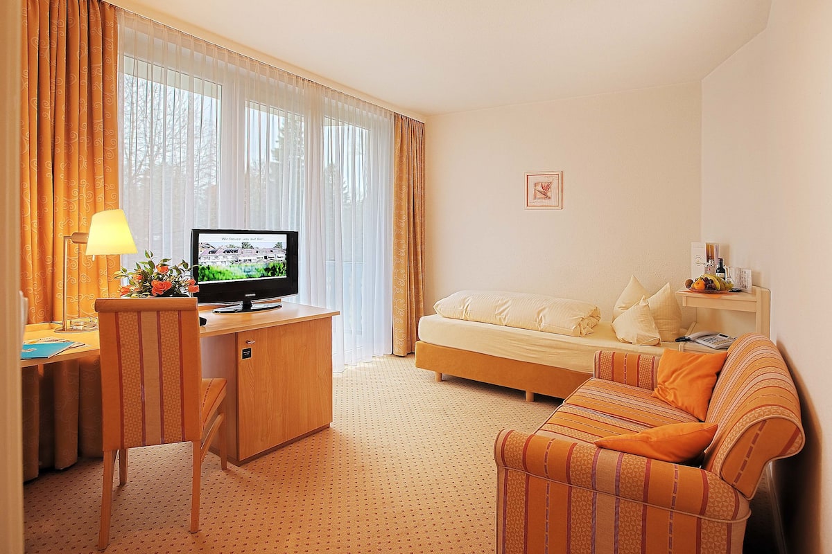 A hotel room with a bed, a sofa, a TV on a wooden desk, and a lamp.