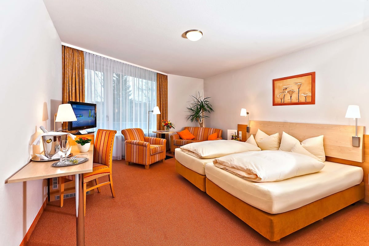 A hotel room with a bed, a desk with a TV, a chair, a small table with a plant, and a window with curtains.