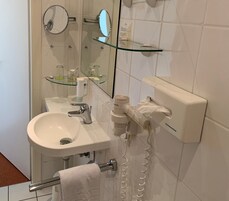Un baño con un lavabo blanco, un espejo, un estante de vidrio con artículos de aseo, un perchero para toallas y un secador de pelo.
