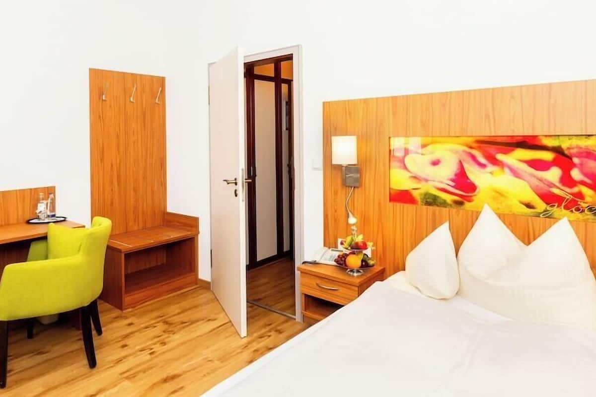 A hotel room with a wooden desk, a bright green chair, a bed with white pillows, a nightstand with a lamp, and a colorful painting on the wall.