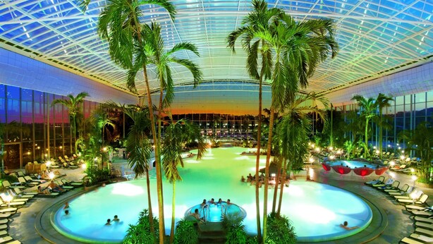 An indoor swimming pool with multiple levels, surrounded by palm trees and lounge chairs. - Hotel Sonnengarten (Bad Woerishofen)