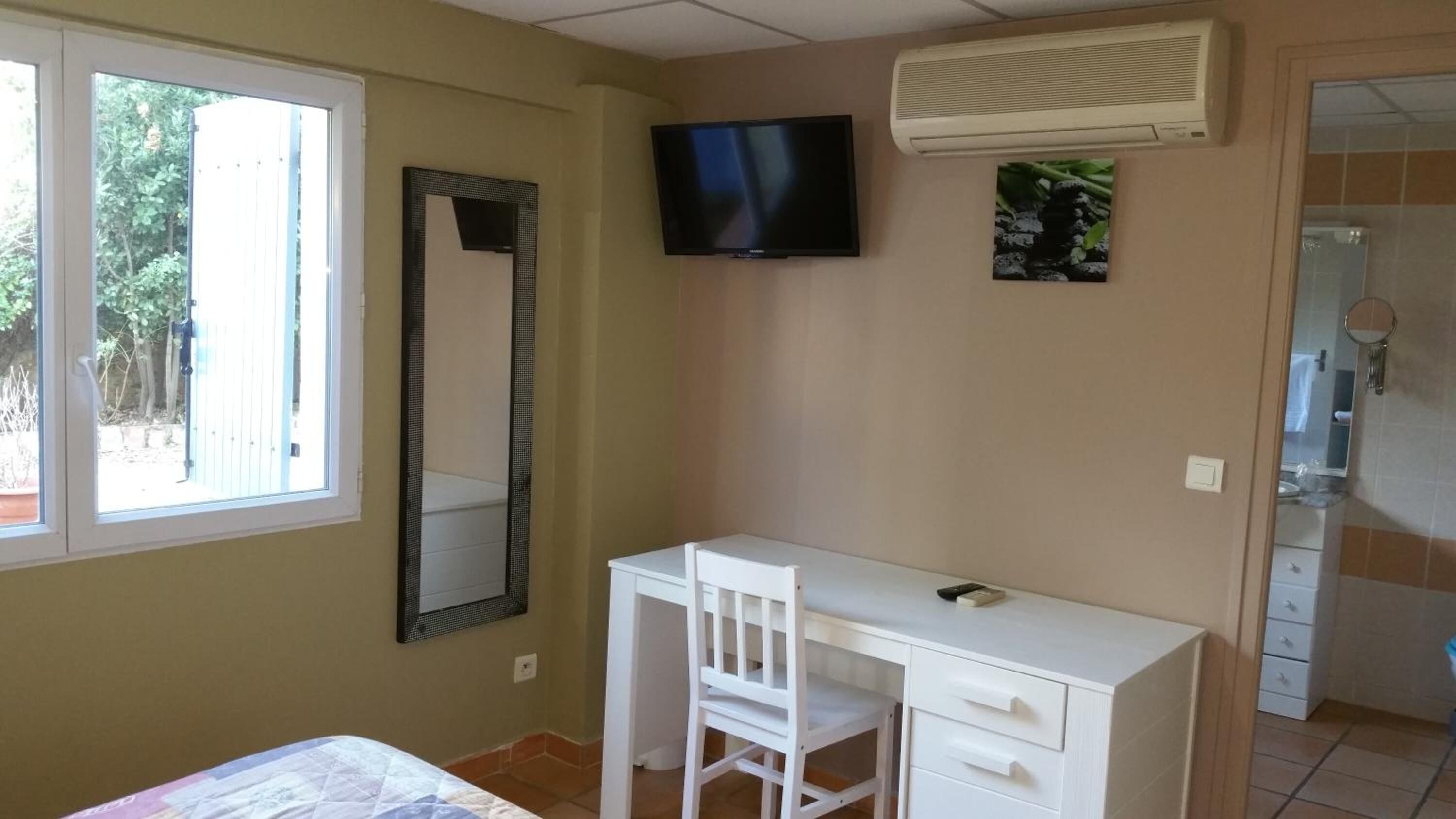 Quarto com cama, escrivaninha, cadeira, televisão e banheiro visível por uma porta aberta.