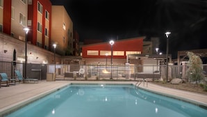 Une piscine entourée d’une clôture, avec des chaises longues, et un bâtiment dont les murs extérieurs sont peints en rouge et en beige, visible au fond.