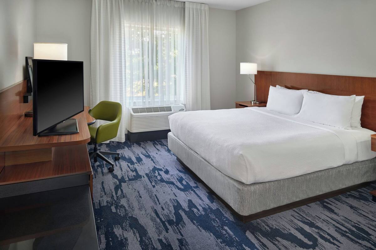 Une chambre d’hôtel avec un grand lit, un bureau doté d’un téléviseur à écran plat, un fauteuil vert et une fenêtre ornée de rideaux légers.
