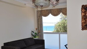 A living room with a dark grey sofa, a ceiling light fixture, a large window with curtains, and a view of the sea. - Hotel Bat Yam (Bat Yam)