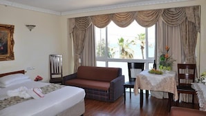 A hotel room with a bed, a sofa, a dining table with a vase of flowers, and a view of palm trees through large windows. - Hotel Bat Yam (Bat Yam)