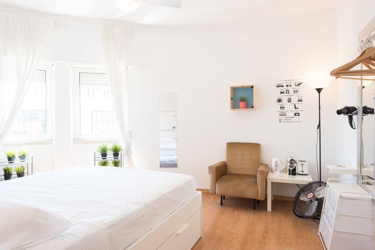A bright, minimalist bedroom with a large bed, a small desk, a chair, and a fan.