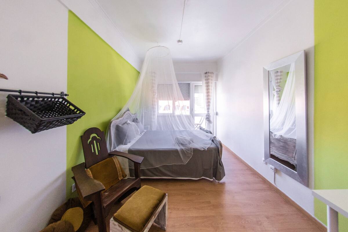 A bedroom with a bed under a mosquito net, a chair, a small table, and a window with curtains.