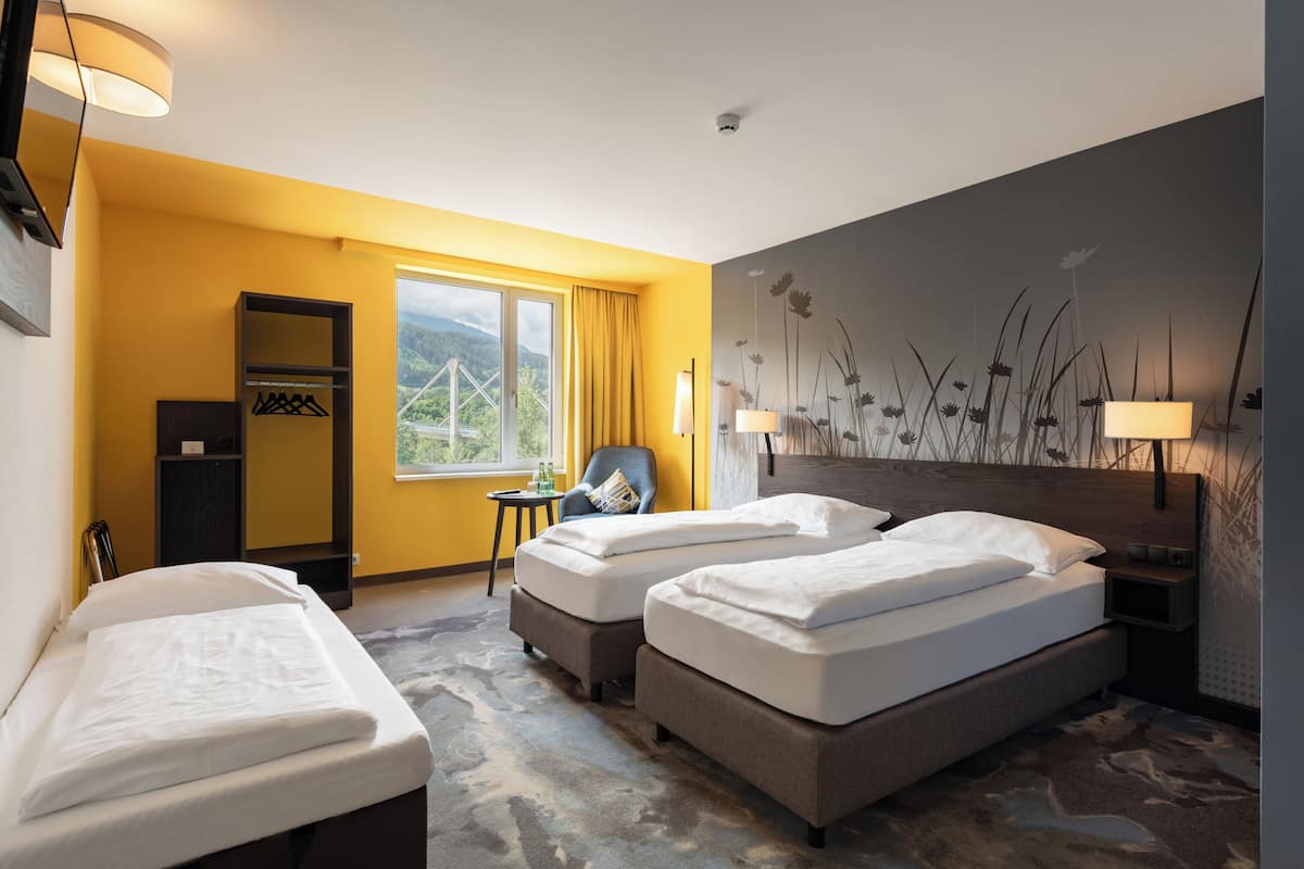 Une chambre d’hôtel avec deux lits simples, une chaise, une petite table, une armoire et une vue sur un pont et la verdure, visible par la fenêtre.