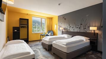 A hotel room with two single beds, a chair, a small table, a wardrobe, and a view of a bridge and greenery through the window.