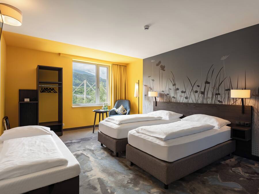 A hotel room with two single beds, a chair, a small table, a wardrobe, and a view of a bridge and greenery through the window.
