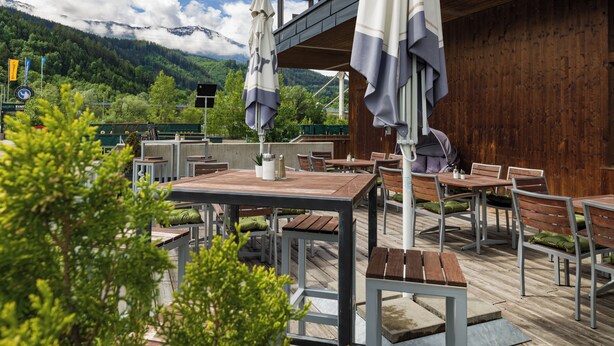 An outdoor patio with wooden tables and chairs, umbrellas, and a view of mountains in the background. - Hwest Hotel Hall (Hall In Tirol)