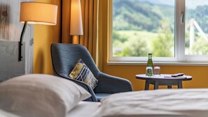 A hotel room with a bed, a chair, a small table with a bottle and glasses, and a window with a view of greenery and mountains. - Hwest Hotel Hall (Hall In Tirol)