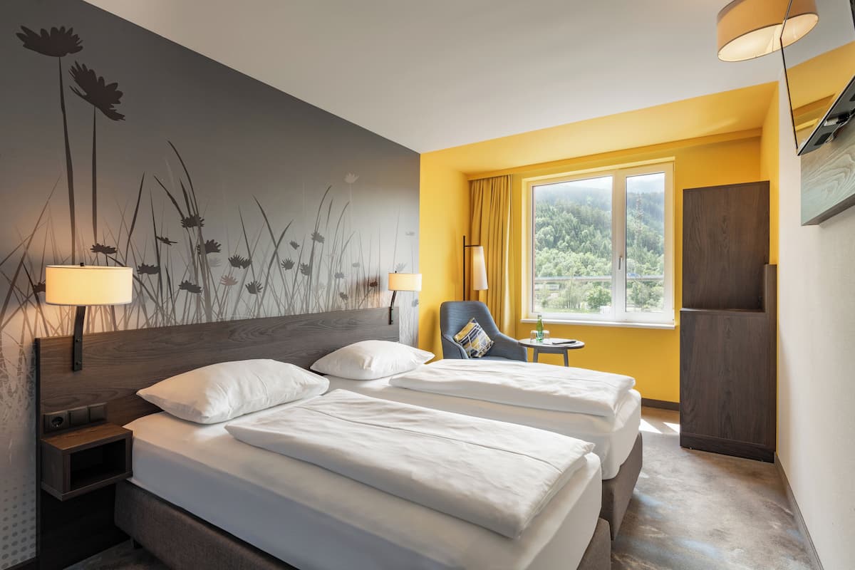 Une chambre d’hôtel avec deux lits, une chaise, une petite table, une fenêtre avec des rideaux et un mur orné d’une fresque florale.