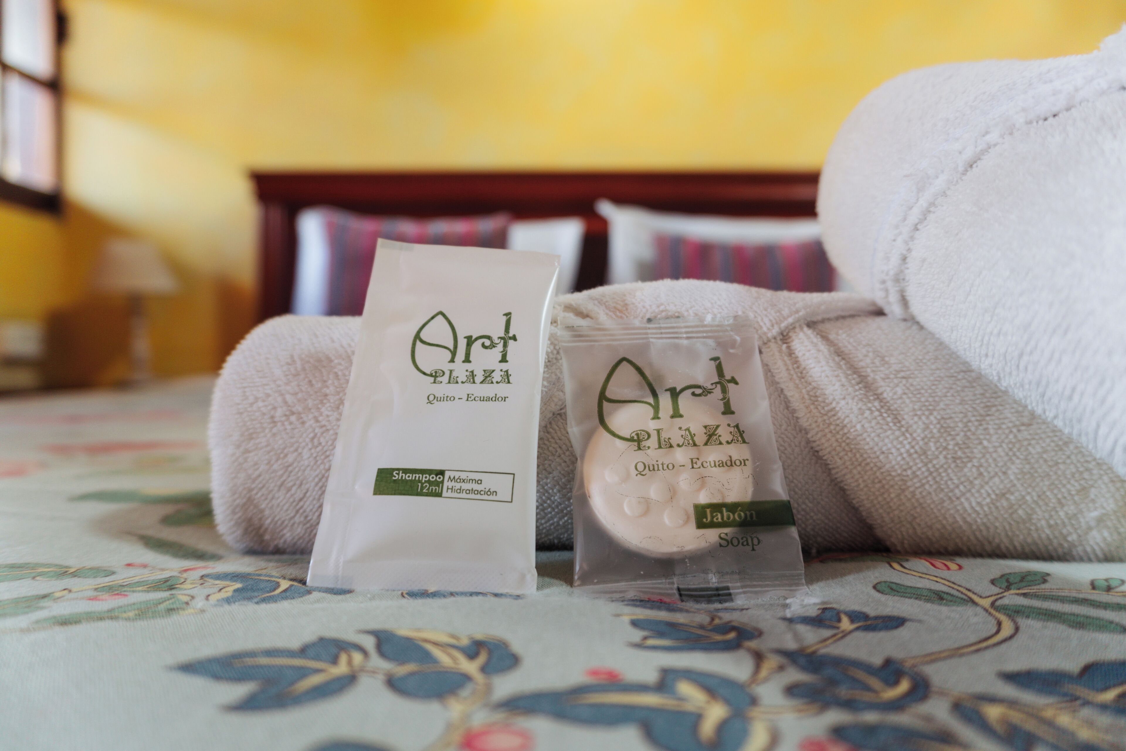 A bed with a floral bedspread, two white towels, and two packages of Art Plaza shampoo and soap.