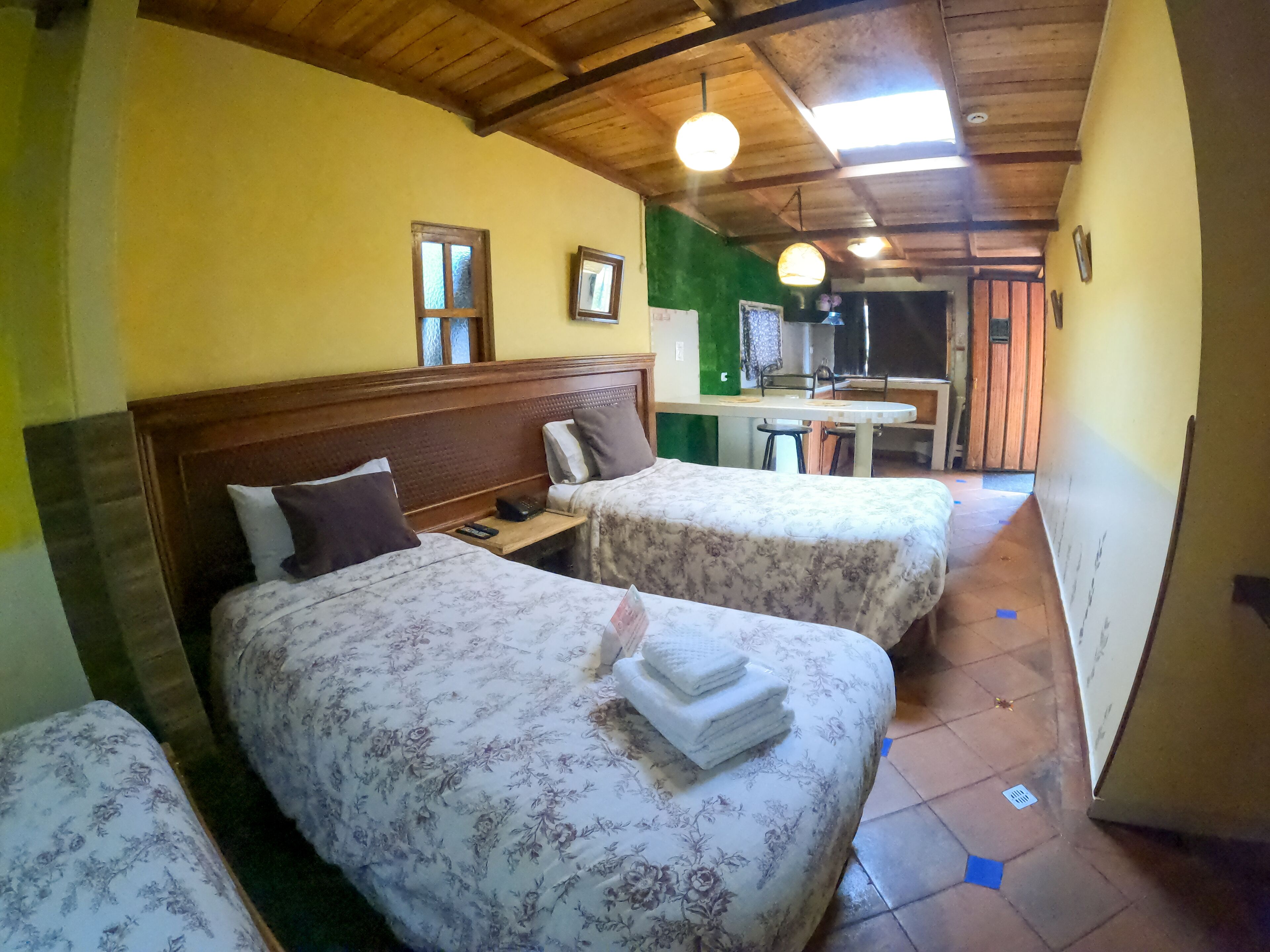 Una habitación con dos camas, techo de madera y una pequeña kitchenette al fondo.
