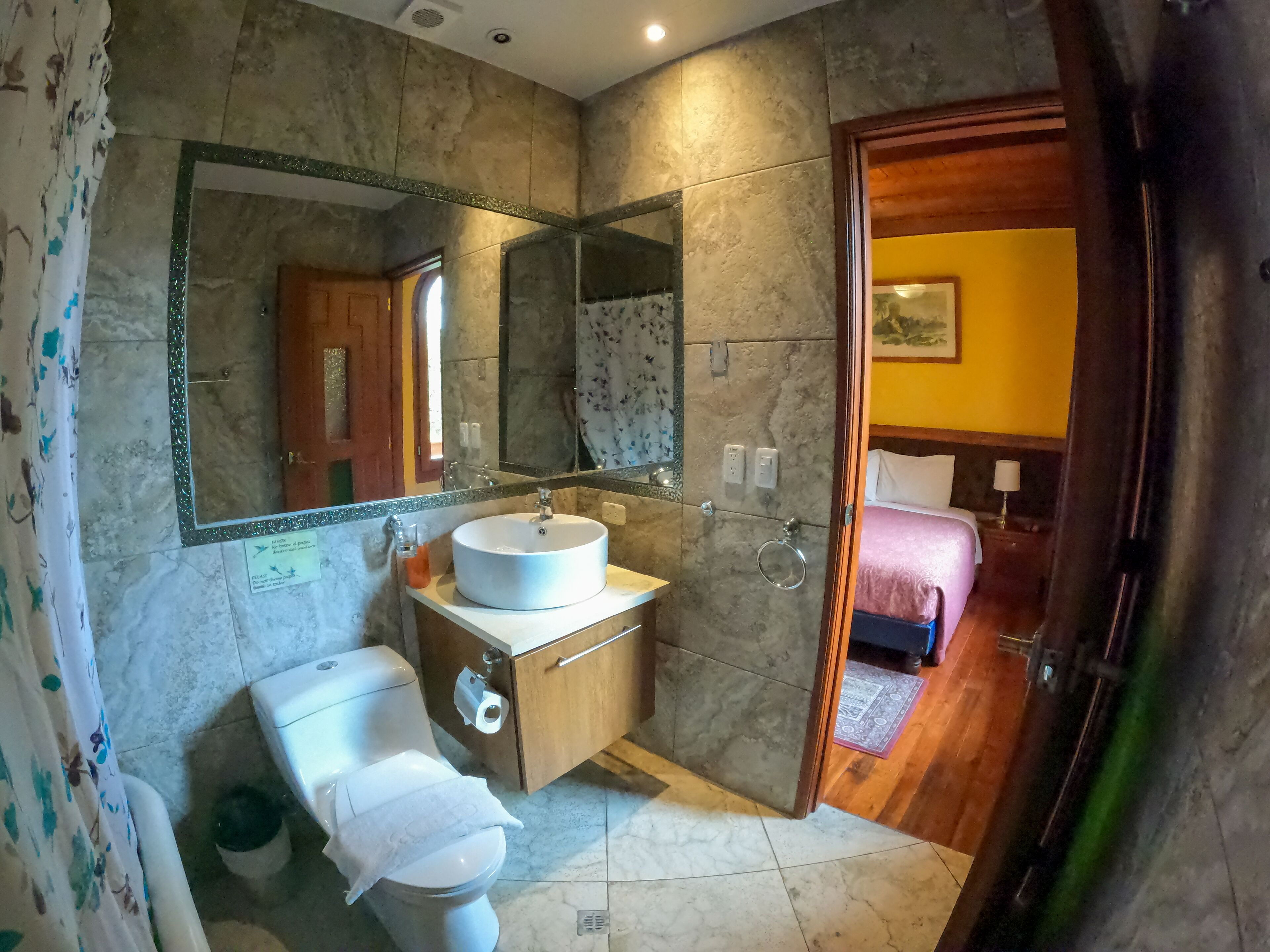 Une salle de bain avec des toilettes, un lavabo et un miroir. Une porte en bois donne sur une chambre à coucher équipée d’un lit et d’une table de chevet.
