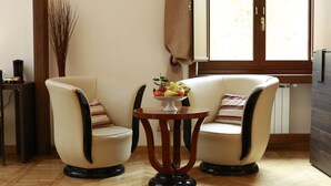 Ein Zimmer mit zwei Sesseln, einem kleinen runden Tisch mit Obstschale, einer hohen Vase mit Zweigen und einem Fenster mit Vorhängen.