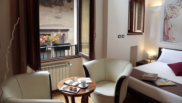 Ein Hotelzimmer mit einem Bett, zwei Sesseln, einem kleinen Tisch mit einem Buch und einer Tasse sowie Blick auf einen Balkon mit Blumen.