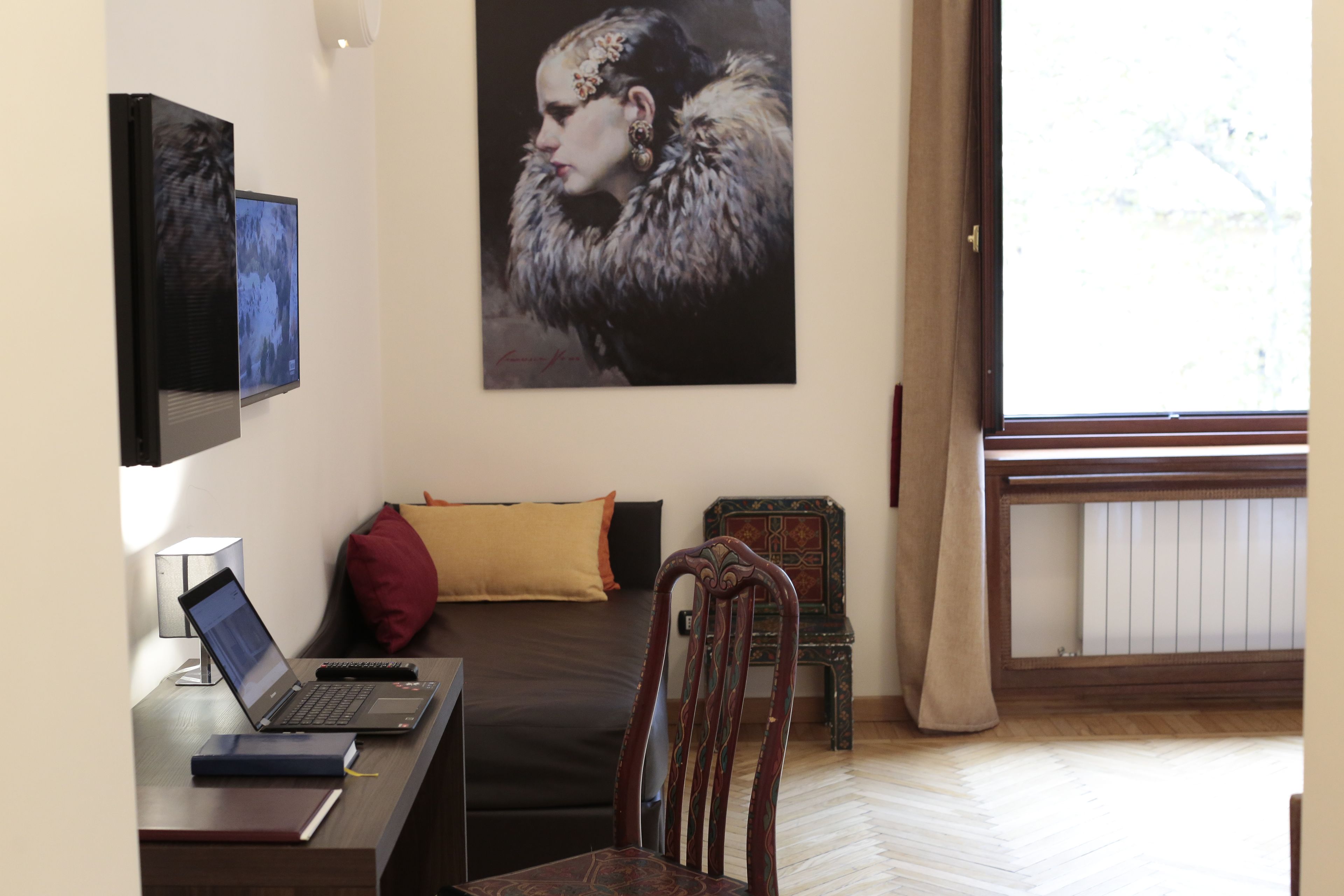A bedroom with a bed, a desk with a laptop, a chair, a small table, a decorative chair, a painting, and a window with curtains.
